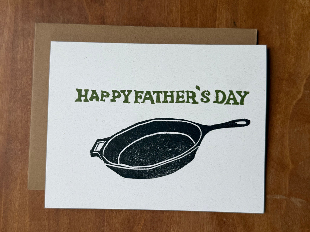 Father's Day card with a cast iron frying pan stamped illustration and 'Happy Father's Day' text on a wooden surface.