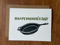 Father's Day card with a cast iron frying pan stamped illustration and 'Happy Father's Day' text on a wooden surface.