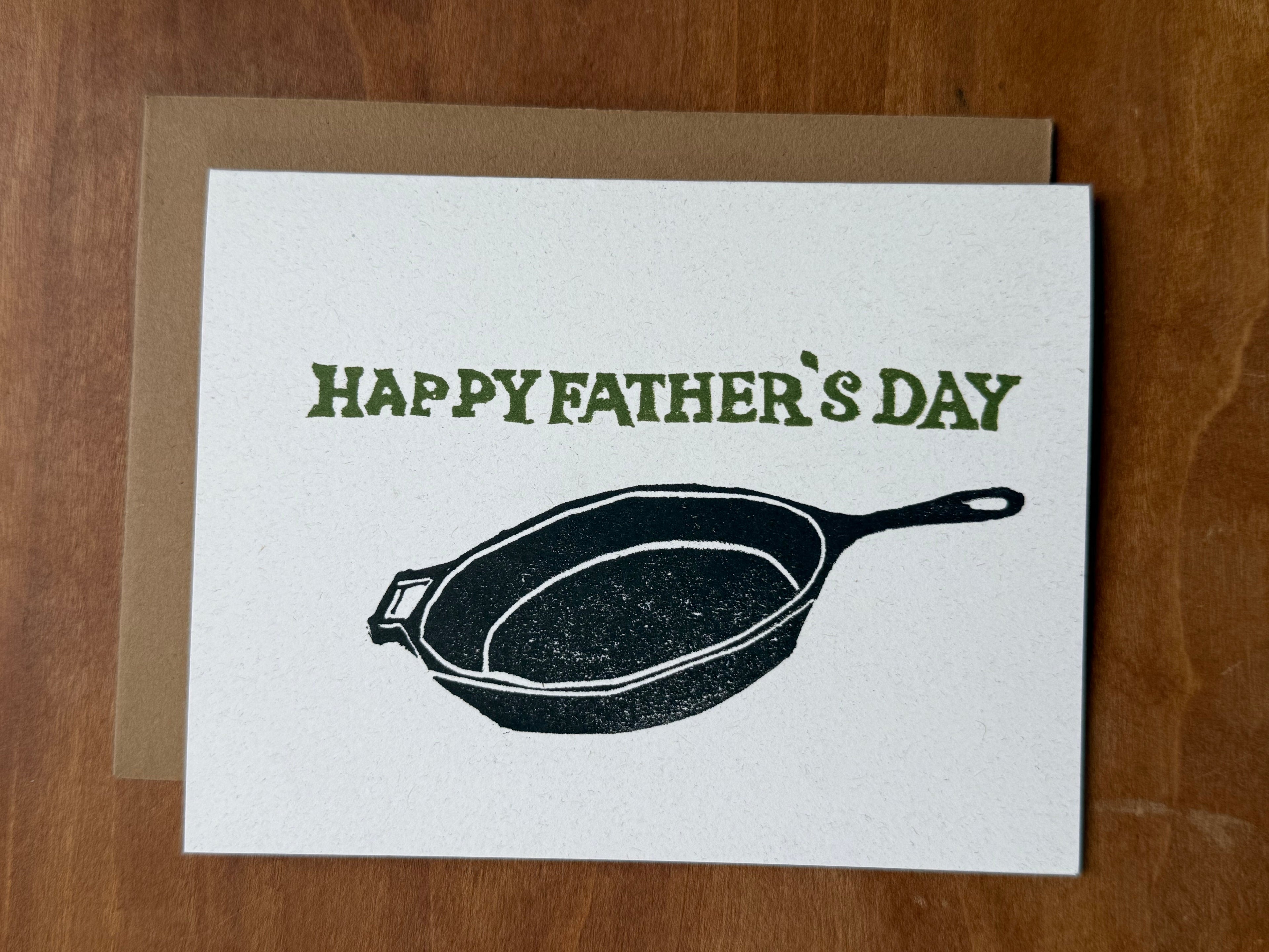 Father's Day card with a cast iron frying pan stamped illustration and 'Happy Father's Day' text on a wooden surface.