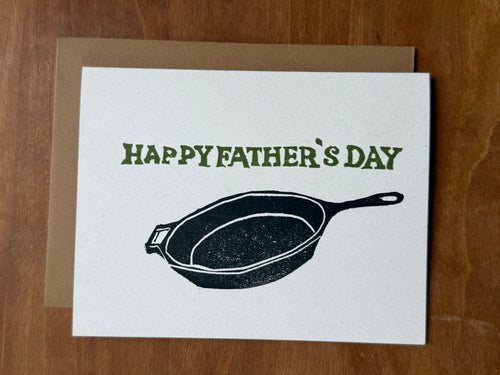 Father's Day card with a cast iron frying pan stamped illustration and 'Happy Father's Day' text on a wooden surface.